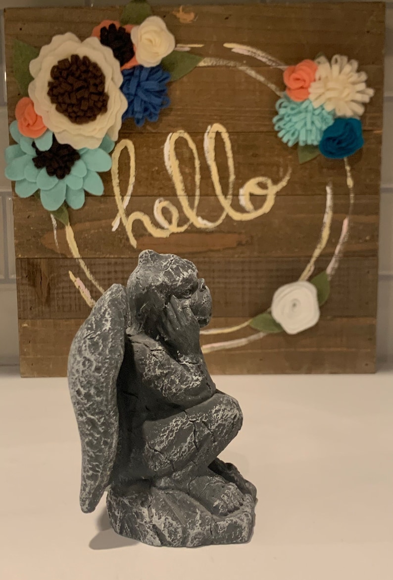 May include: A gray stone gargoyle statue with wings, sitting on a white surface. The gargoyle is looking to the right. The background is a wooden sign with the word "hello" written in yellow paint and decorated with felt flowers.