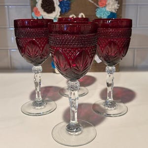 Pode incluir: Um conjunto de cinco taças de vinho de cristal vermelho rubi. Cada taça apresenta um padrão detalhado na taça e uma haste clara e ornamentada. As taças são exibidas sobre uma superfície branca, com um painel decorativo de madeira no fundo.