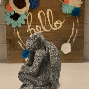 May include: A gray stone gargoyle statue with wings, sitting on a base. The gargoyle is looking to the left and has a textured surface. The statue is in front of a wooden sign with the word "hello" written in yellow paint.
