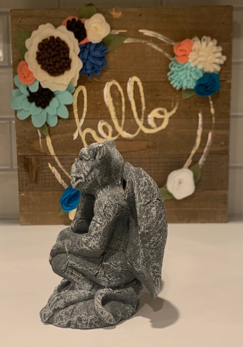 May include: A gray stone gargoyle statue with wings, sitting on a base. The gargoyle is looking to the left and has a textured surface. The statue is in front of a wooden sign with the word "hello" written in yellow paint.