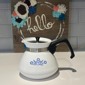May include: White enamel coffee pot with a black handle and lid. The pot has a silver band around the middle and a blue floral design. Behind the pot is a wooden sign with the word "hello" and a floral wreath.