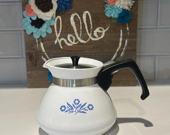 Op de afbeelding: Witte emaille koffiepot met een zwarte handgreep en deksel. De pot heeft een zilveren band en een blauw bloemmotief. Achter de pot staat een houten bord met het woord "hello" en een bloemenkrans.