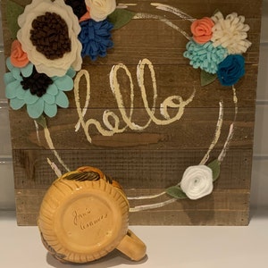 May include: A yellow ceramic mug with brown and black painted details. The mug has the words "Jan's Ceramics" written on the bottom. The mug is sitting in front of a wooden sign with the word "hello" written in white paint. The sign is decorated with felt flowers in shades of white, blue, and pink.