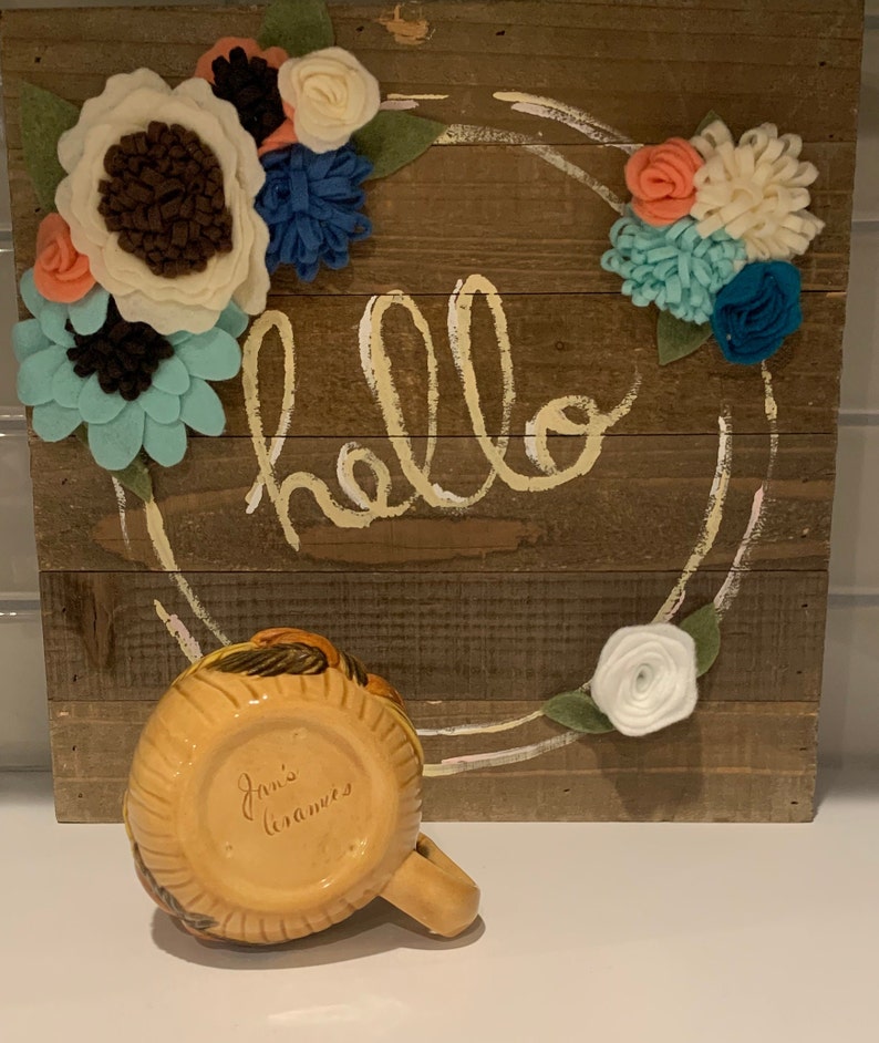 May include: A yellow ceramic mug with brown and black painted details. The mug has the words "Jan's Ceramics" written on the bottom. The mug is sitting in front of a wooden sign with the word "hello" written in white paint. The sign is decorated with felt flowers in shades of white, blue, and pink.