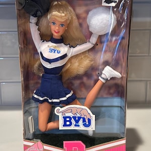 May include: A Special Edition University Barbie doll in a clear plastic box. The doll is dressed as a cheerleader in a white and navy blue uniform with the BYU logo. The box has pink and white accents and the words "University Barbie".