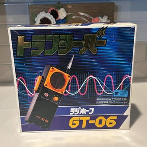 May include: A rectangular product box for a GT-06 transceiver featuring a black and orange handheld radio illustration against a blue background with white and red wave patterns. The box displays Japanese text, a pilot lamp, and volume dial.
