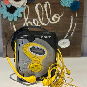 May include: A vintage Sony Sports radio with yellow accents and matching earbuds. The radio features buttons for FM, AM, and TV, with the word "SONY" prominently displayed. The background includes a wooden sign with the word "hello" and a floral wreath.