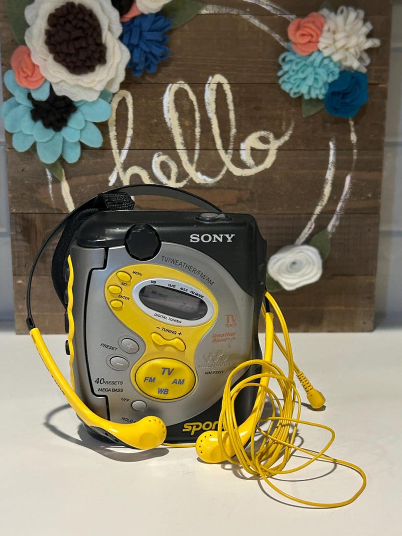 May include: A vintage Sony Sports radio with yellow accents and matching earbuds. The radio features buttons for FM, AM, and TV, with the word "SONY" prominently displayed. The background includes a wooden sign with the word "hello" and a floral wreath.
