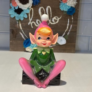 May include: A ceramic elf figurine with blonde hair, a pink hat with a white pom-pom, a green tunic with a gold heart, and pink legs, sitting on a dark brown log-shaped base. The elf has pointed ears, blue eyes, and a smiling expression.