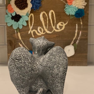 May include: A gray stone sculpture of two cats with wings, sitting back to back. The sculpture is in front of a wooden sign with the word "hello" written on it. The sign is decorated with felt flowers in shades of blue, white, and pink.