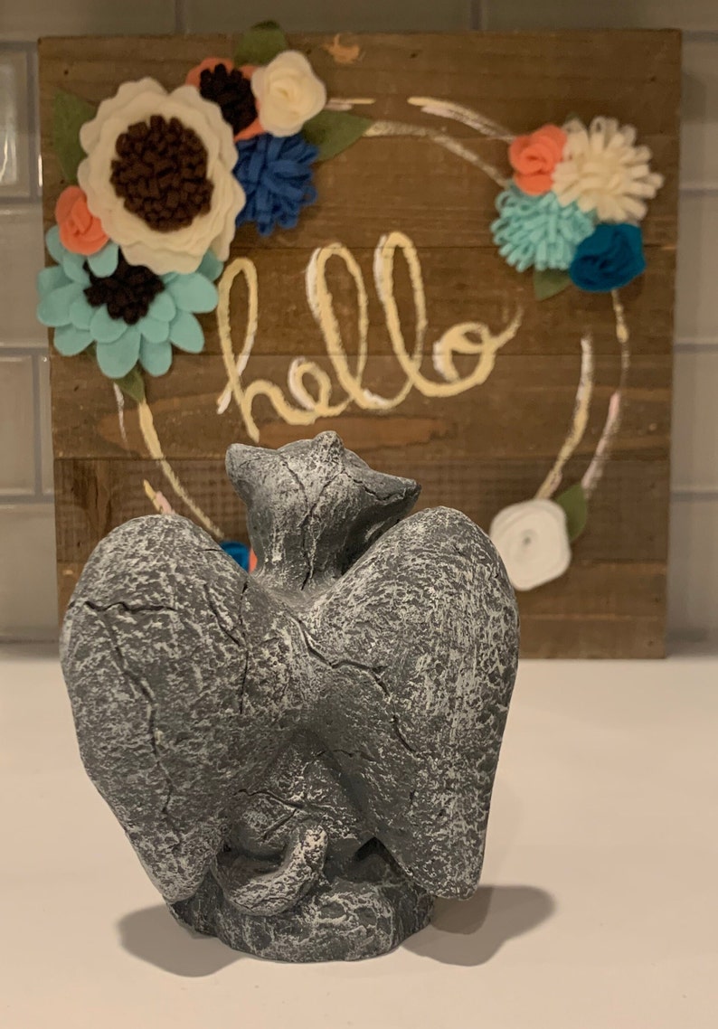 May include: A gray stone sculpture of two cats with wings, sitting back to back. The sculpture is in front of a wooden sign with the word "hello" written on it. The sign is decorated with felt flowers in shades of blue, white, and pink.