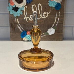 Pode incluir: Pote de vidro âmbar com tampa e rolha decorativa. O pote é redondo com uma tampa a condizer. A rolha é alongada e tem uma pega transparente. No fundo, uma placa de madeira com a palavra "hello" e detalhes florais.