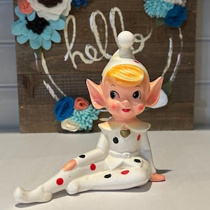 Peut inclure: Une figurine d'elfe en céramique en position assise, vêtue d'une tenue blanche ornée de pois noirs et rouges. L'elfe a des cheveux blonds, des oreilles pointues et un chapeau blanc assorti avec un pompon. Un détail en forme de cœur doré est sur le col.
