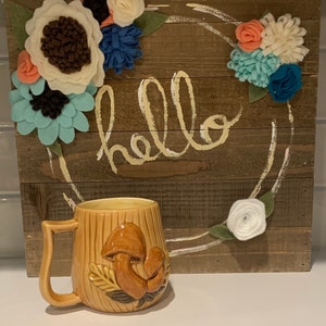 May include: A brown ceramic mug with a mushroom design. The mug has a handle and is sitting on a white surface. The background is a wooden sign with the word "hello" written in white paint and a wreath of felt flowers.