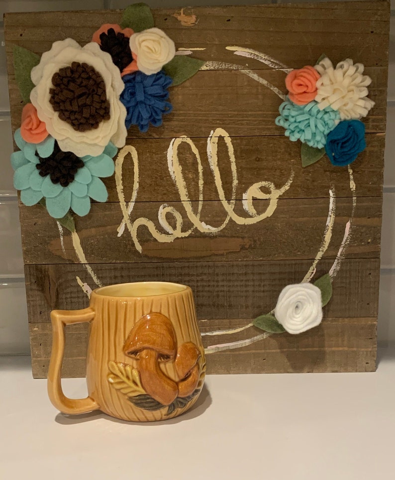 May include: A brown ceramic mug with a mushroom design. The mug has a handle and is sitting on a white surface. The background is a wooden sign with the word "hello" written in white paint and a wreath of felt flowers.
