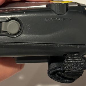 May include: Close-up of a black Sony Walkman. The device has a gray button, a volume control, and a black wrist strap. The top of the device has a yellow accent.