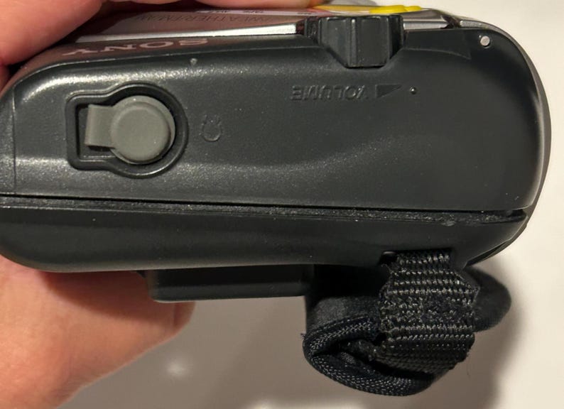 May include: Close-up of a black Sony Walkman. The device has a gray button, a volume control, and a black wrist strap. The top of the device has a yellow accent.