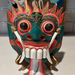 Puede incluir: Una máscara de madera tallada a mano con una cara verde, detalles rojos y dorados. La máscara presenta grandes ojos negros, dientes blancos y una lengua roja y blanca. Es un objeto decorativo.