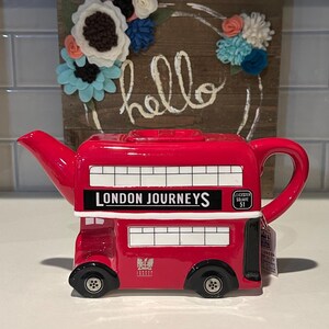 Puede incluir: Tetera de cerámica roja con forma de autobús de dos pisos londinense. Tiene ruedas negras, ventanas blancas y la inscripción "LONDON JOURNEYS". Una etiqueta cuelga del asa. Un letrero de madera con "hello" y adornos florales en el fondo.