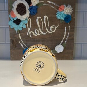 May include: A ceramic mug with a cream-colored base and a handle with black polka dots. The mug features a colorful animal print design. The bottom of the mug has a Disney Parks stamp. A wooden sign with the word "hello" and floral accents is in the background.