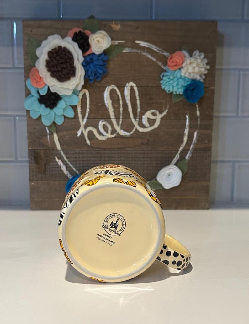 May include: A ceramic mug with a cream-colored base and a handle with black polka dots. The mug features a colorful animal print design. The bottom of the mug has a Disney Parks stamp. A wooden sign with the word "hello" and floral accents is in the background.
