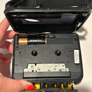 May include: Black portable cassette player with the top open, revealing the interior components. A Duracell battery is visible. The player has a black strap and a serial number 5069237.