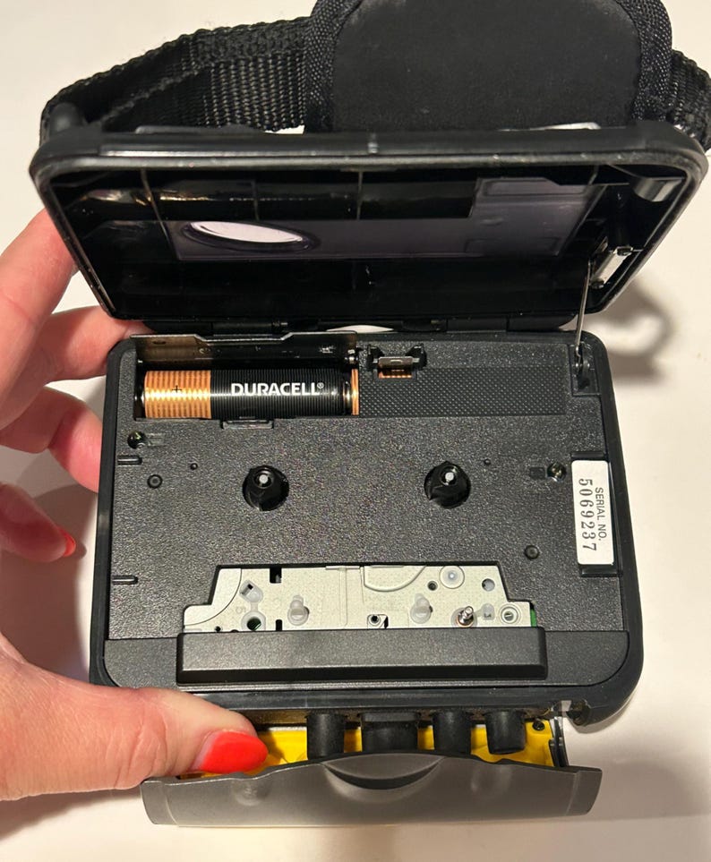 May include: Black portable cassette player with the top open, revealing the interior components. A Duracell battery is visible. The player has a black strap and a serial number 5069237.