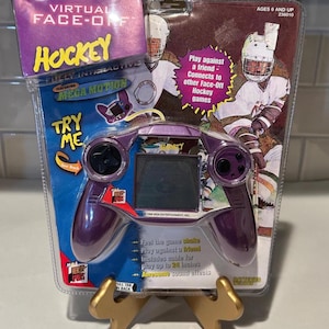 May include: A purple and black handheld "Virtual Face-Off Hockey" game in its original packaging. The game features a screen, two control pads, and the text "TRY ME". The packaging includes hockey player graphics and the text "AGES 6 AND UP".