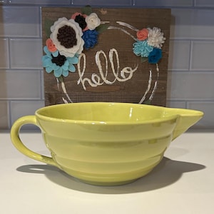 May include: A yellow ceramic mixing bowl with a handle and a spout. The bowl has horizontal ridges and sits in front of a wooden sign with the word "hello" and felt flower decorations.