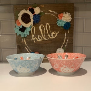 May include: Two ceramic bowls with a cat design. The bowl on the left is light blue with a white cat and the bowl on the right is pink with a white cat. Both bowls have a speckled glaze.