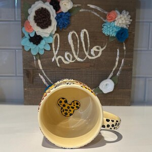 May include: A ceramic coffee mug with a yellow interior and a black and yellow spotted exterior. The handle is also spotted. The interior features a Mickey Mouse head design. Behind the mug is a wooden sign with the word "hello" and a floral wreath.