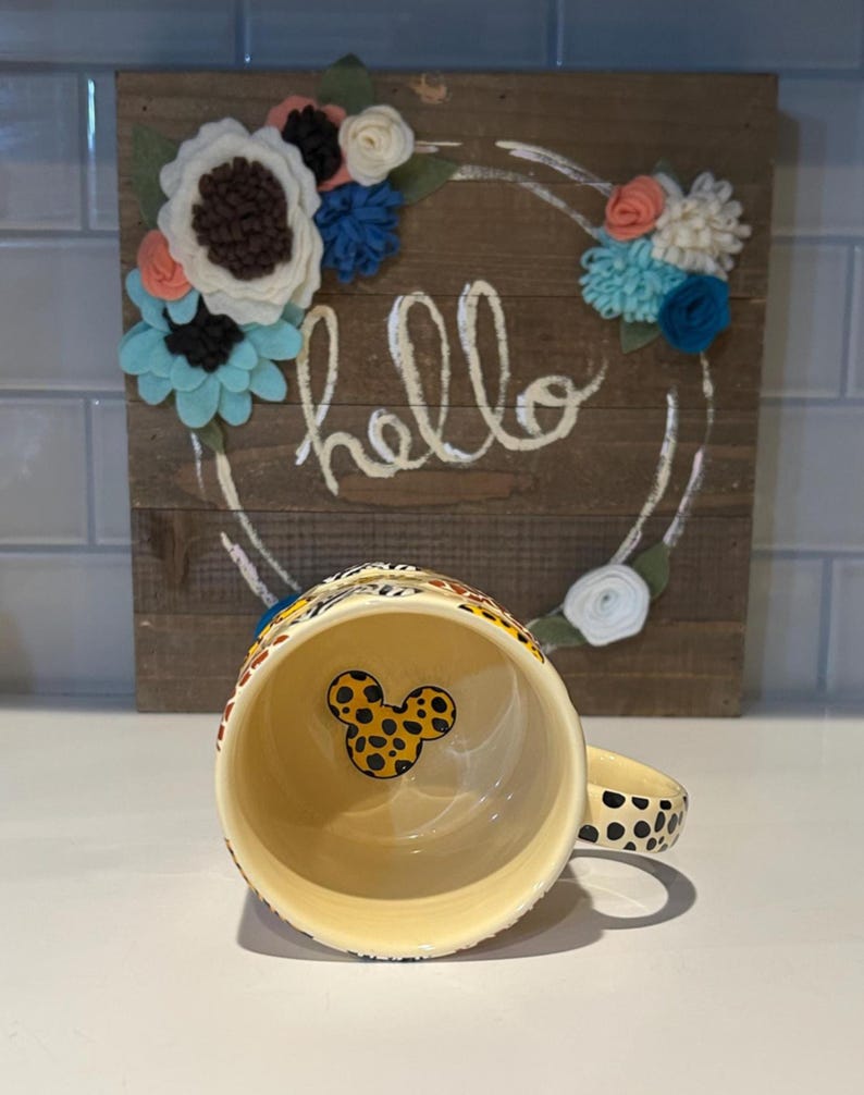 May include: A ceramic coffee mug with a yellow interior and a black and yellow spotted exterior. The handle is also spotted. The interior features a Mickey Mouse head design. Behind the mug is a wooden sign with the word "hello" and a floral wreath.