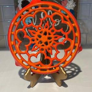 May include: A circular cast iron trivet featuring a vibrant orange and red gradient finish with an intricate openwork floral pattern. The trivet is displayed upright on a gold-toned metal stand, showcasing its detailed lattice design.