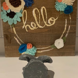May include: A gray concrete bunny figurine with a rustic wooden sign behind it. The sign has a wreath of felt flowers in shades of blue, white, and coral. The sign says "hello" in a yellow script font.
