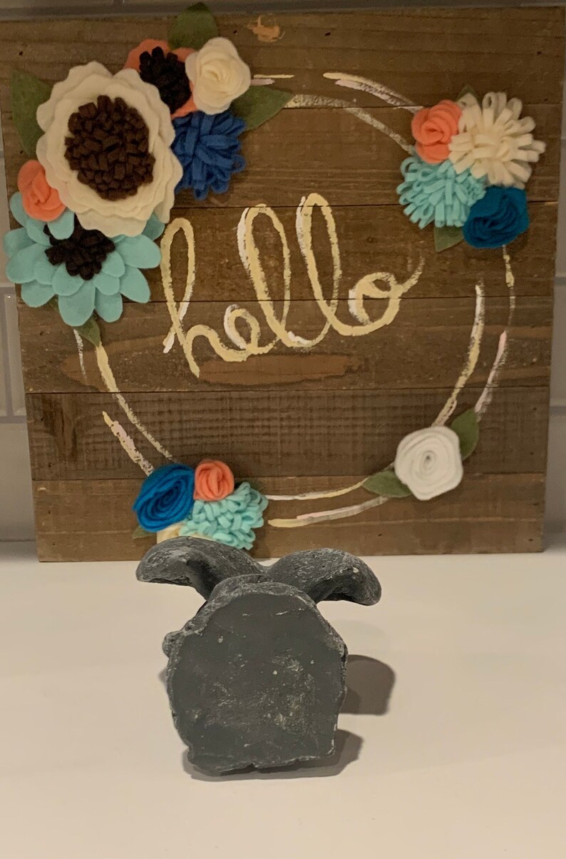 May include: A gray concrete bunny figurine with a rustic wooden sign behind it. The sign has a wreath of felt flowers in shades of blue, white, and coral. The sign says "hello" in a yellow script font.