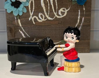 Puede incluir: Una figura de piano negro con un personaje de dibujos animados sentado con un vestido rojo, tocando el piano. La figura está frente a un letrero de madera con la palabra "hello" y flores de fieltro.
