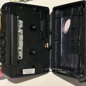 May include: Black portable cassette player with the door open. The player has a black body with a yellow side. The open door reveals the tape compartment. A black strap is attached to the side. The serial number 5069237 is visible.