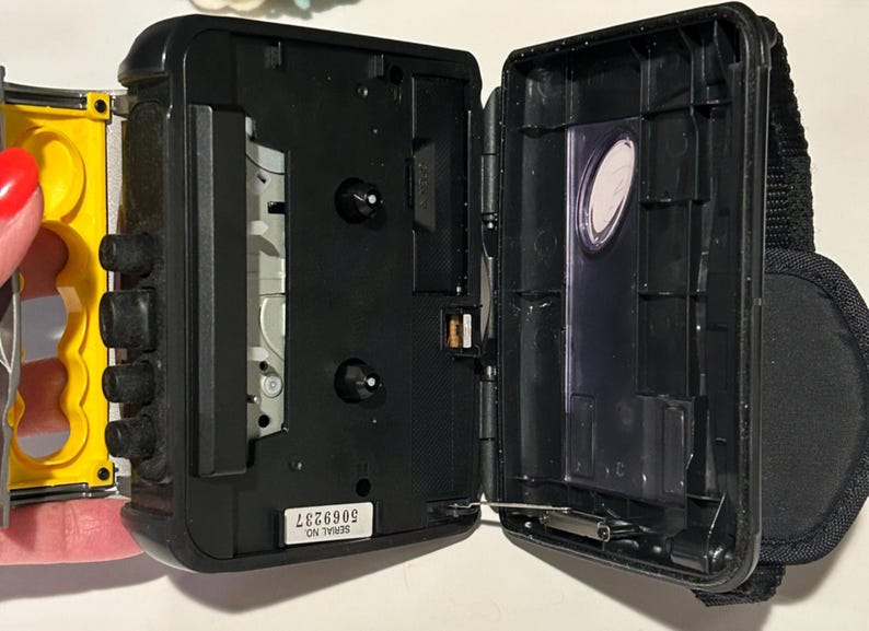 May include: Black portable cassette player with the door open. The player has a black body with a yellow side. The open door reveals the tape compartment. A black strap is attached to the side. The serial number 5069237 is visible.