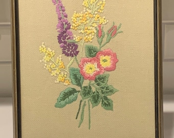 Op de afbeelding: Geborduurde bloemenkunst in een rechthoekige lijst. Het boeket heeft roze, gele en paarse bloemen met groene bladeren op een beige achtergrond. De ingelijste kunst wordt tentoongesteld op een goudkleurig ezel.