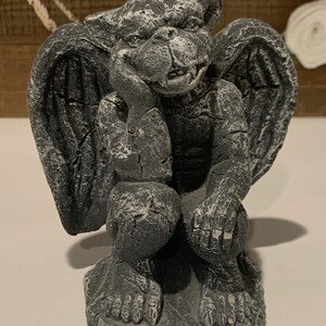 May include: A gray stone gargoyle statue with wings, sitting with its head resting on its hand. The gargoyle has a detailed, textured surface and a menacing expression.