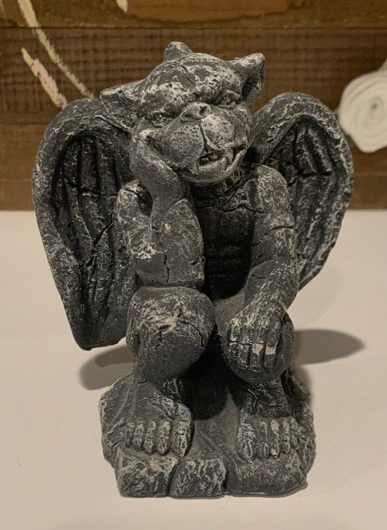 May include: A gray stone gargoyle statue with wings, sitting with its head resting on its hand. The gargoyle has a detailed, textured surface and a menacing expression.