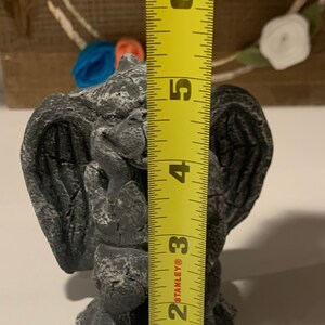 May include: A gray stone gargoyle statue with wings, measuring approximately 5 inches tall. The statue is sitting on a white surface next to a yellow measuring tape.