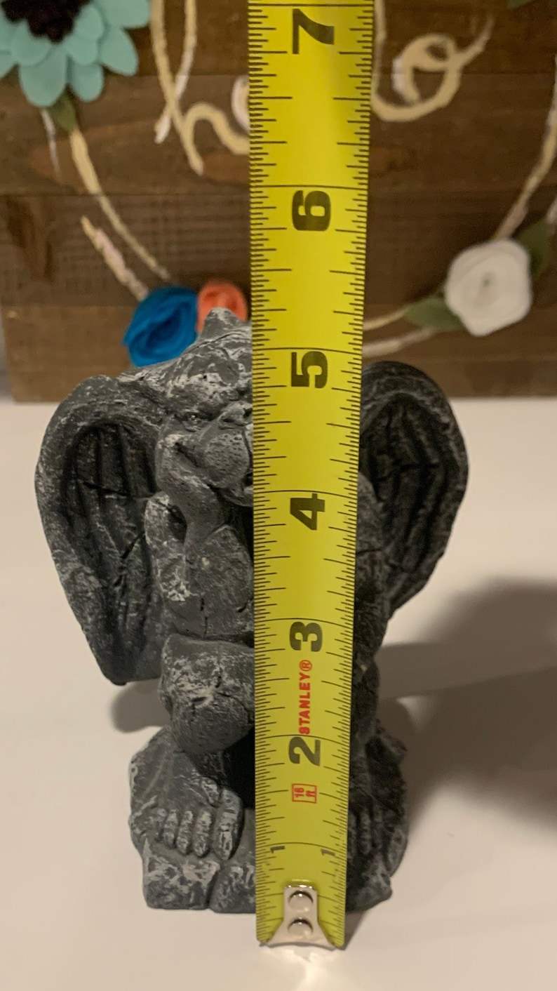 May include: A gray stone gargoyle statue with wings, measuring approximately 5 inches tall. The statue is sitting on a white surface next to a yellow measuring tape.