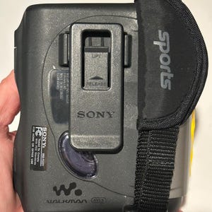 May include: A gray and black Sony Walkman WM-FS221 with a black "Sports" strap. The device has a clip and the Sony logo. The text "WALKMAN" and "AVLS" are visible on the device.