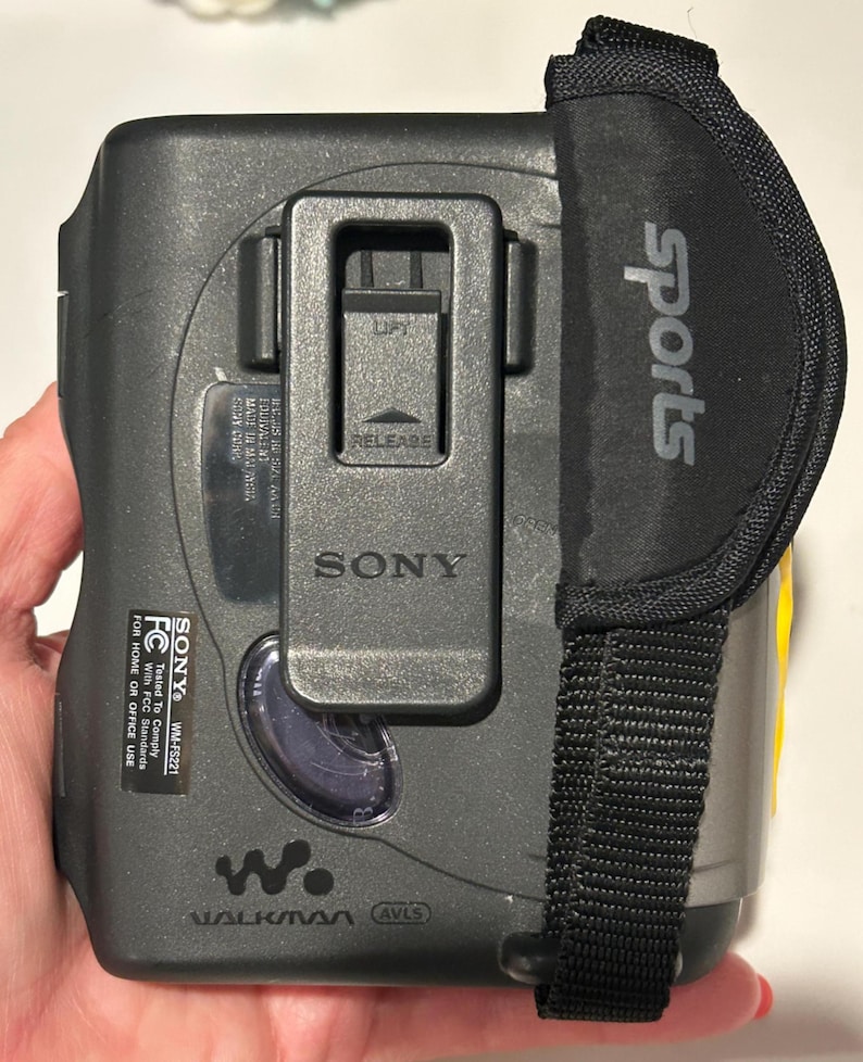 May include: A gray and black Sony Walkman WM-FS221 with a black "Sports" strap. The device has a clip and the Sony logo. The text "WALKMAN" and "AVLS" are visible on the device.