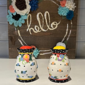 May include: A set of two ceramic salt and pepper shakers with a white background and a floral pattern. The shakers have black polka dots, red and yellow accents, and a black and white trim.