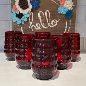 May include: Set of six red glass tumblers with a bubbled texture. The glasses are arranged in a row on a white surface.