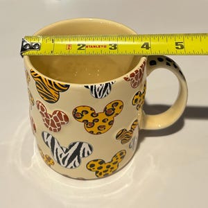 May include: Cream-colored ceramic mug with Mickey Mouse head shapes in various animal print patterns, including zebra, leopard, and tiger. The mug is approximately 3.5 inches in diameter and has a matching handle.