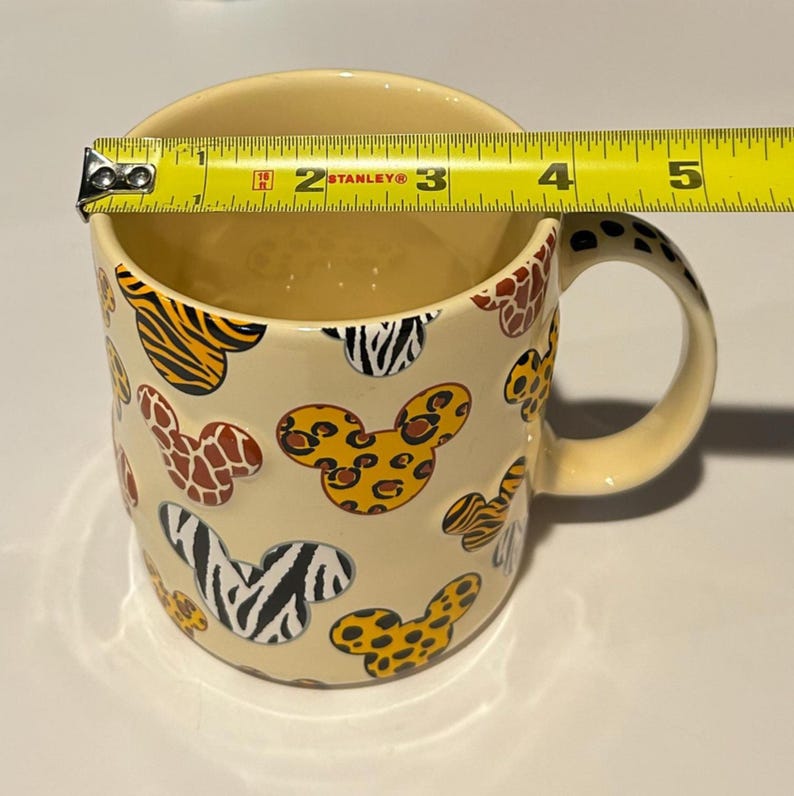 May include: Cream-colored ceramic mug with Mickey Mouse head shapes in various animal print patterns, including zebra, leopard, and tiger. The mug is approximately 3.5 inches in diameter and has a matching handle.