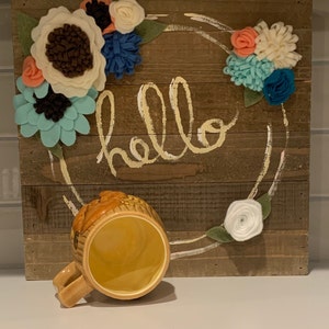 May include: A wooden sign with the word "hello" painted in yellow on it. The sign is decorated with a wreath of felt flowers in shades of white, blue, and pink. A yellow ceramic mug with a brown design is sitting in front of the sign.